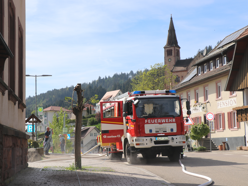 Ein rotes Feuerwehrfahrzeug steht in einem Dorf auf der Straße, während ein Feuerwehrschlauch auf den Gehweg ausgerollt ist.