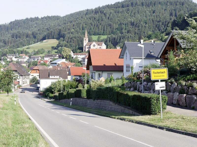 Eine malerische Straße führt durch ein Dorf mit einer Kirche im Hintergrund, umgeben von grünen Hügeln und verschiedenen Häusern.