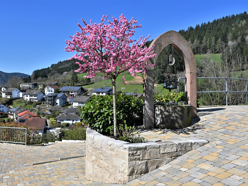 Eine malerische Landschaft zeigt einen blühenden Baum in einem steinernen Pflanzbeet, umgeben von einem Bogen mit einer Glocke, vor einem Dorf in hügeliger, bewaldeter Umgebung.