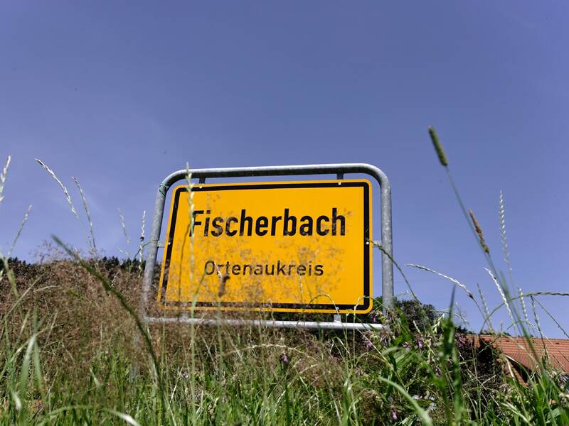 Ein gelbes Ortseingangsschild mit der Aufschrift "Fischerbach, Ortenaukreis" steht inmitten von hohem Gras vor einem blauen Himmel.