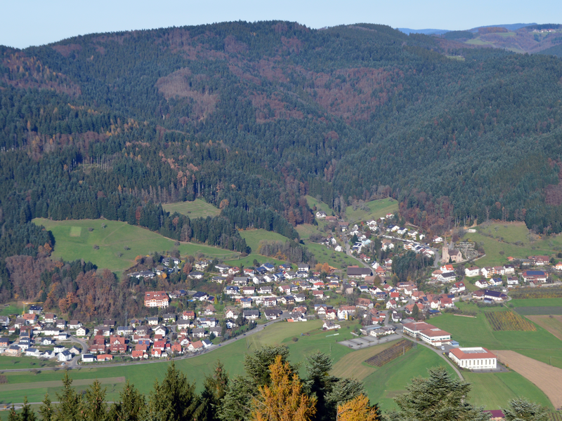 Eine kleine Stadt liegt eingebettet in einer von bewaldeten Hügeln umgebenen Landschaft mit Feldern und vereinzelten Gebäuden.