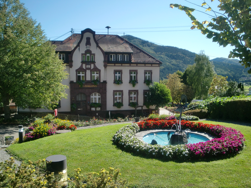 Ein großes, historisches Gebäude mit der Aufschrift "Altes Schulhaus" steht vor einer von Blumen umgebenen Springbrunnenanlage in einer grünen, bergigen Landschaft.