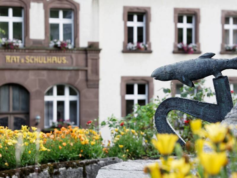 Im Vordergrund steht eine Skulptur eines Wals mit einem Anker, umgeben von gelben und orangefarbenen Blumen, während im Hintergrund die Fassade eines Gebäudes mit der Aufschrift "Kath. Schulhaus" zu sehen ist.