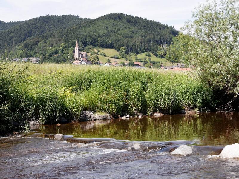 Ein ruhiger Fluss fließt im Vordergrund durch eine grüne Landschaft mit einem kleinen Dorf und einer Kirche im Hintergrund, umgeben von bewaldeten Hügeln.