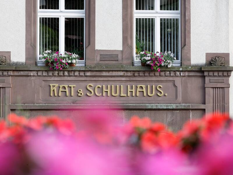 Ein altes Gebäude mit der Aufschrift "RAT & SCHULHAUS" über dem Eingang und Blumen in Fensternähe.