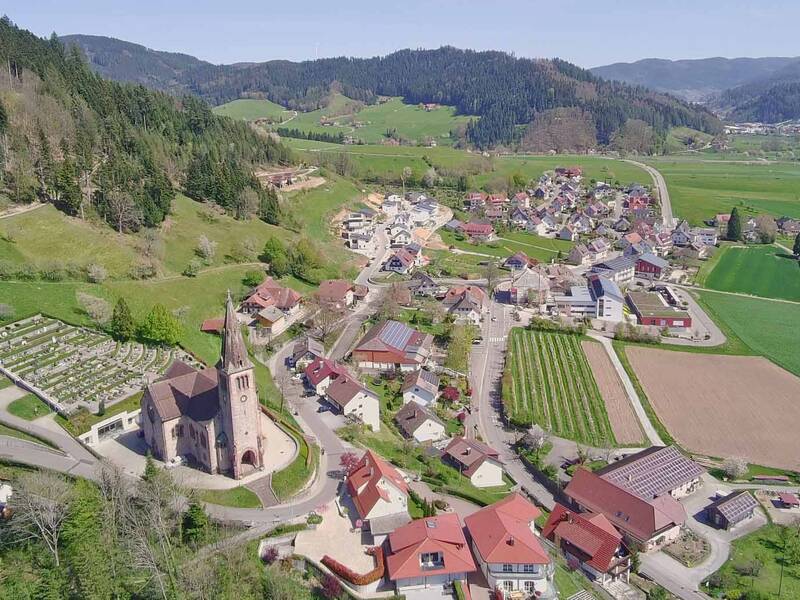 Luftaufnahme eines Dorfes in einer ländlichen Gegend mit einer Kirche im Vordergrund, umgeben von Feldern, Wiesen und Hügeln.