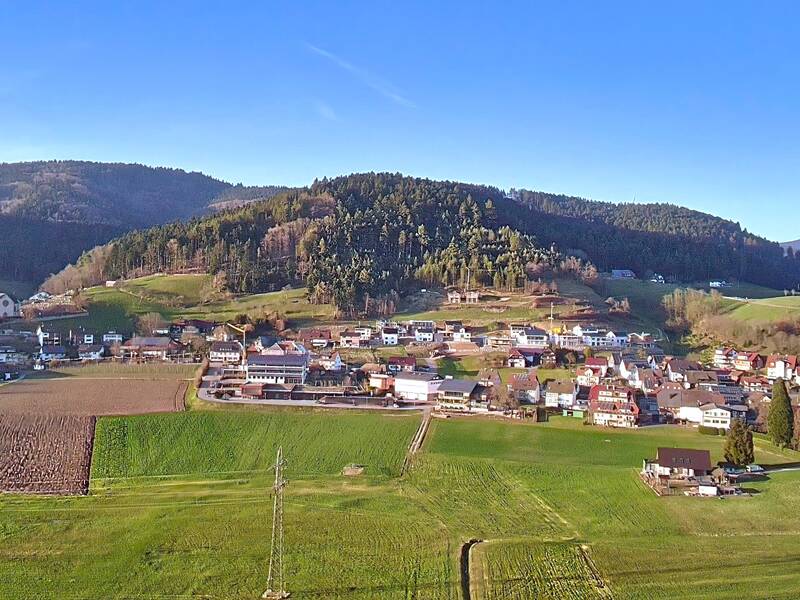 Die Luftaufnahme zeigt ein malerisches Dorf mit verstreuten Häusern und einer Kirche, umgeben von grünen Feldern und bewaldeten Hügeln unter einem klaren, blauen Himmel.