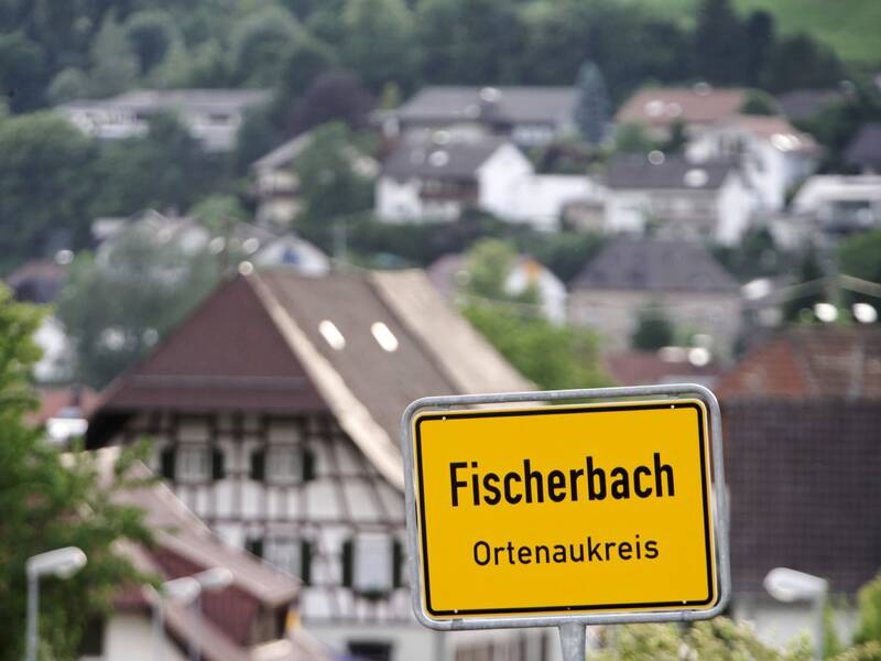 Im Vordergrund ist ein gelbes Ortsschild mit der Aufschrift "Fischerbach Ortenaukreis" zu sehen, im Hintergrund erstreckt sich eine unscharfe, grüne Landschaft mit Häusern.