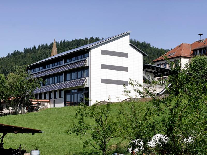 Ein modernes Gebäude mit Solarpaneelen steht auf einem grünen Hügel vor einem Hintergrund aus Wald und einem Kirchturm.