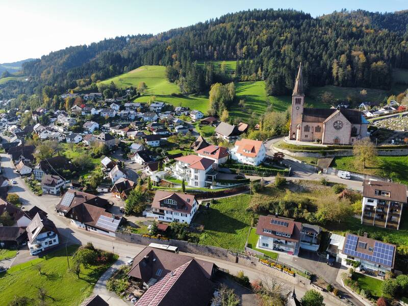 Das Bild zeigt eine malerische Dorfansicht mit vielen Häusern, einer großen Kirche und bewaldeten Hügeln im Hintergrund.