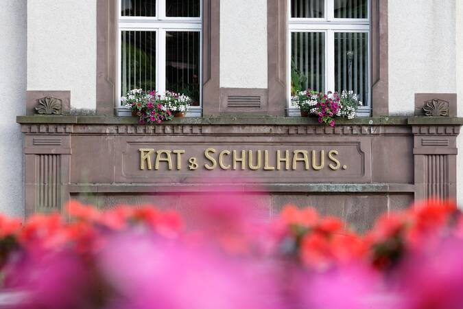 Die Fassade eines Gebäudes mit der Aufschrift "Rat & Schulhaus" und blühenden Blumen vor den Fenstern.