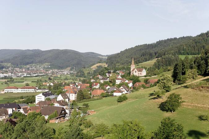 Eine Panoramaaufnahme zeigt ein malerisches Dorf inmitten grüner Hügel mit einer Kirche, die aus den traditionellen Häusern hervorragt, umgeben von üppigen Wäldern und einem klaren Himmel.