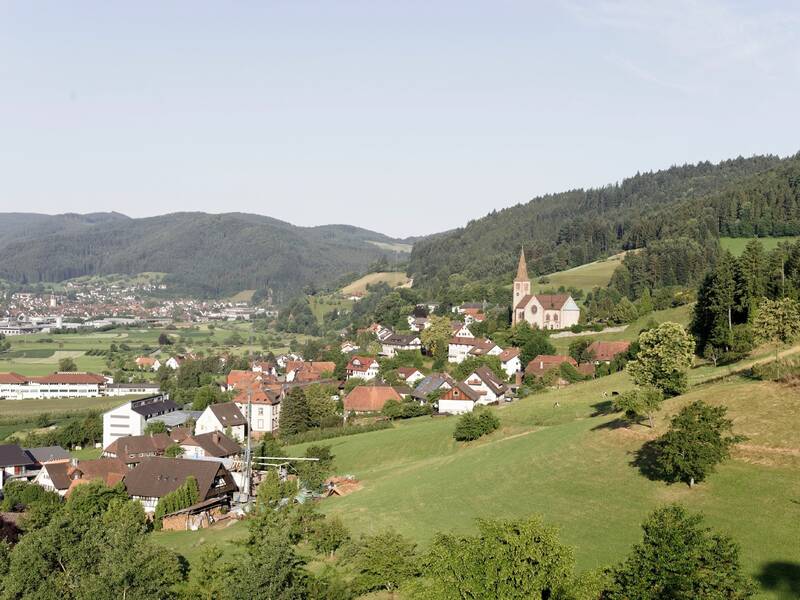 Eine Panoramaaufnahme zeigt ein malerisches Dorf inmitten grüner Hügel mit einer Kirche, die aus den traditionellen Häusern hervorragt, umgeben von üppigen Wäldern und einem klaren Himmel.