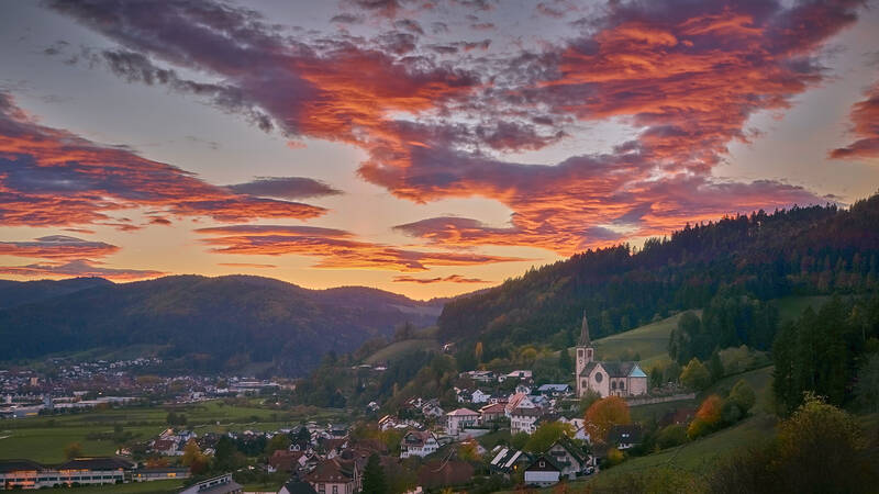 Eine malerische Landschaft zeigt ein Dorf mit einer Kirche im Vordergrund, umgeben von grünen Hügeln, unter einem dramatischen, rot-orangefarbenen Abendhimmel.