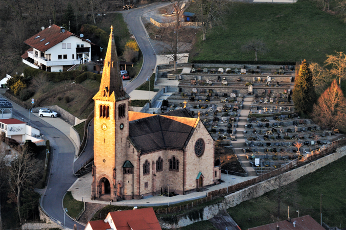 Das Bild zeigt eine Kirche mit einem hohen Turm neben einem Friedhof, umgeben von einer Straße und Häusern in einer hügeligen Landschaft.