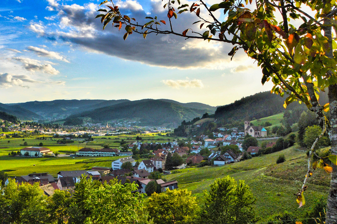 Ein malerisches Dorf mit einer Kirche liegt inmitten grüner Hügel und Wiesen unter einem teils bewölkten Himmel.