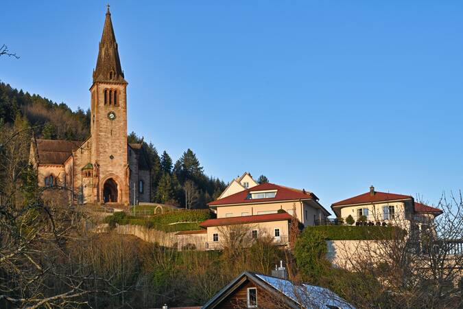 Eine Kirche mit einem hohen Glockenturm steht auf einem Hügel neben mehreren modernen Häusern, umgeben von Bäumen.