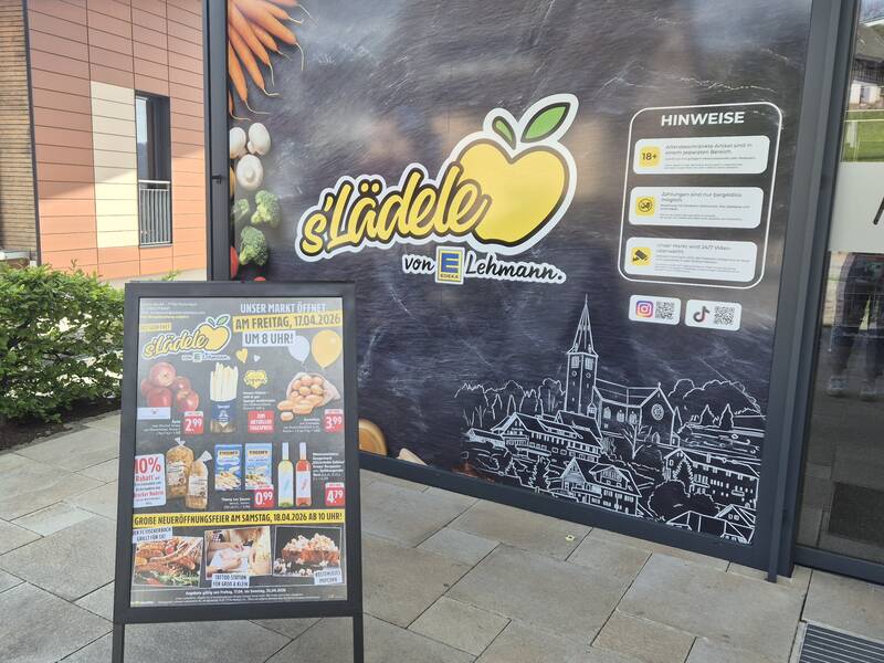 Ein Werbeplakat mit Logos und Informationen zu einem Markt namens "s'Lädle von Lehmann" steht vor einem Geschäft mit großen Schaufenstern, auf denen ein stilisiertes Apfel-Logo und weitere Hinweise zu sehen sind.
