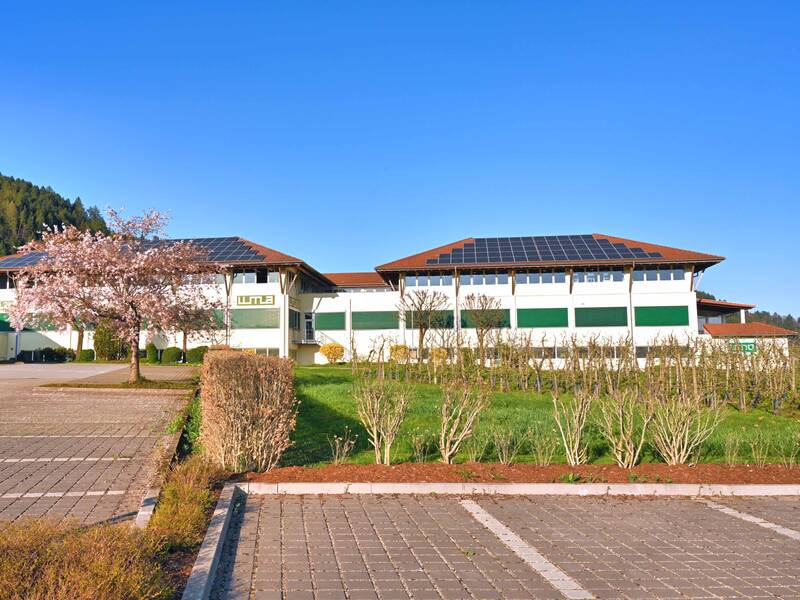 Ein modernes Gebäude mit Solarpanelen auf dem Dach steht vor einem klaren blauen Himmel, umgeben von Bäumen und einem großen Parkplatz.