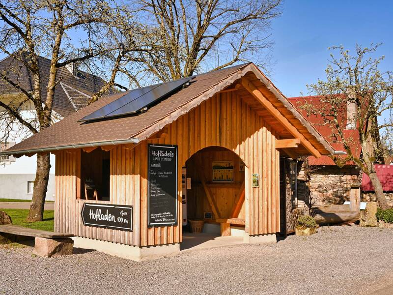 Ein kleiner, rustikaler Holzkiosk mit einem Solarpanel auf dem Dach und Schildern, die auf einen Hofladen hinweisen, steht neben Bäumen und einer steinernen Bank.
