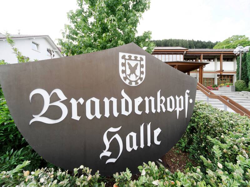 Ein schwarzes Schild mit der Aufschrift "Brandenkopf-Halle" steht vor einem Gebäude, umgeben von grünen Büschen und Bäumen.