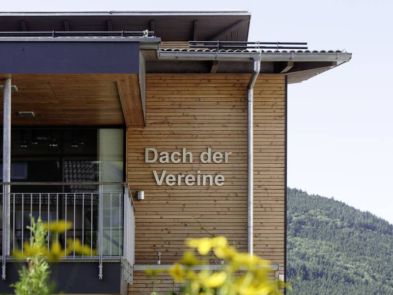 Das Bild zeigt ein modernes Gebäude mit einer Fassade aus Holzlatten. Auf der Holzfassade ist in großen silberfarbenen Buchstaben der Schriftzug „Dach der Vereine“ zu lesen. Das Gebäude verfügt über einen überdachten Balkon oder Eingang mit einer Metallbrüstung und Glasfläche. Die Dachkonstruktion wirkt zeitgemäß, mit einer leichten Neigung und Regenrinnen aus Metall. Im Hintergrund ist ein grüner, bewaldeter Hügel oder Berg zu sehen, was auf eine ländliche oder naturnahe Umgebung hinweist. Im Vordergrund sind unscharfe Pflanzen oder Blumen zu erkennen, die das Bild zusätzlich beleben.