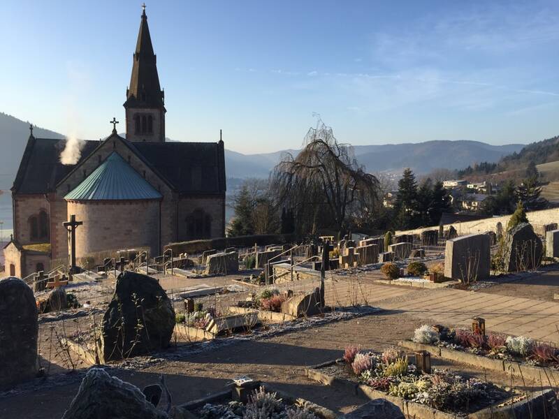 Ein Friedhof liegt vor einer Kirche mit einem Turm, umgeben von einer hügeligen Landschaft.