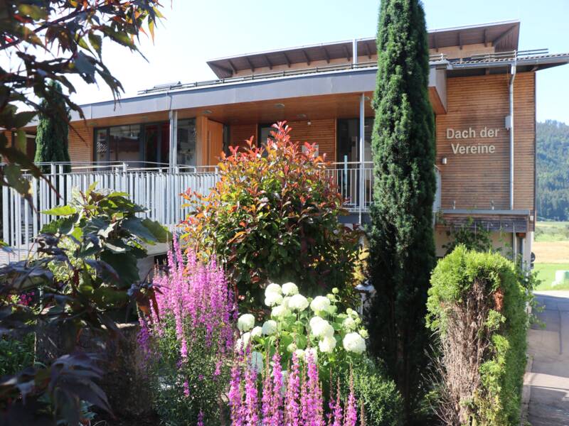 Ein modernes Gebäude mit der Aufschrift "Dach der Vereine" steht hinter einem üppigen Garten mit bunten Blumen und Bäumen.