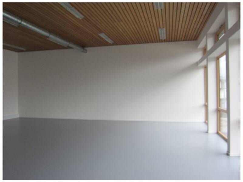 Ein leerer Raum mit grauem Boden, weißen Wänden, einer Holzdecke und großen Fenstern auf der rechten Seite.