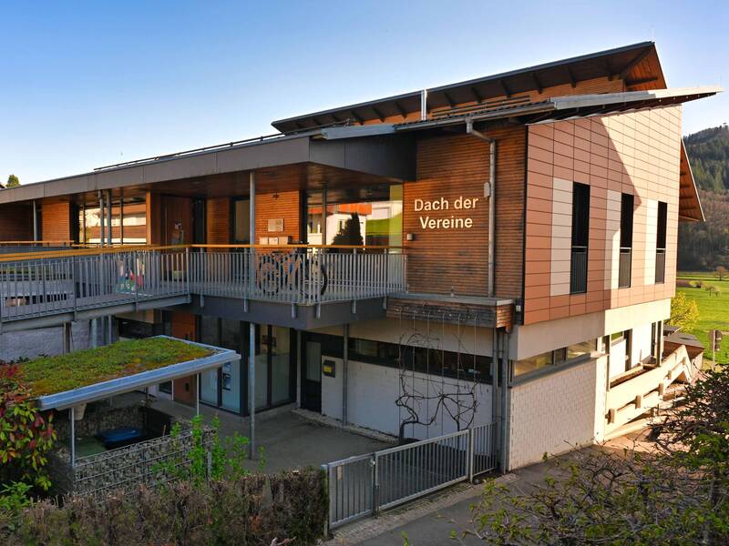 Ein modernes Gebäude mit Holzverkleidung und der Aufschrift "Dach der Vereine" ist von grüner Landschaft umgeben.