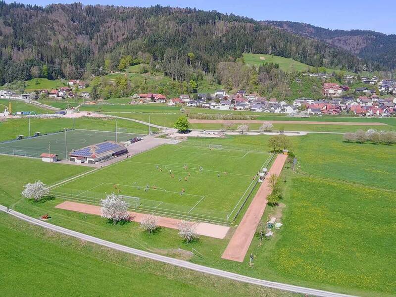 Ein Landschaftsbild zeigt einen Sportplatz mit umliegenden grünen Wiesen, einer kleinen Ortschaft im Hintergrund und bewaldeten Hügeln.