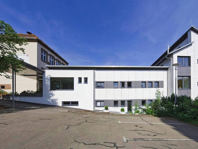 Ein modernes, mehrstöckiges Gebäude mit weißer Fassade steht neben einem älteren, traditionelleren Haus, umgeben von Bäumen und einem Parkplatz.